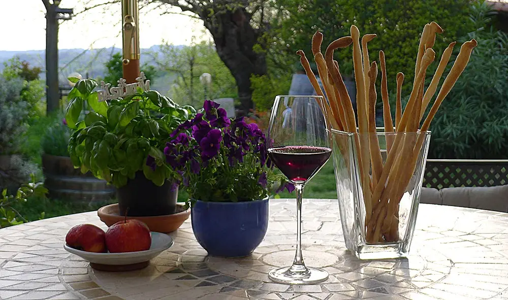 Ein hohes Glas mit dünnen, gedrehten Grissini auf einem Gartentisch, daneben ein Glas Rotwein, ein Basilikumtopf, Stiefmütterchen und eine Schale Äpfel, im Hintergrund eine Hügellandschaft