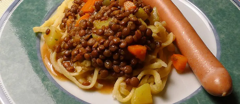 Ein Teller mit Spätzle, braunen Linsen mit Karottenwürfeln und einem Wienerle